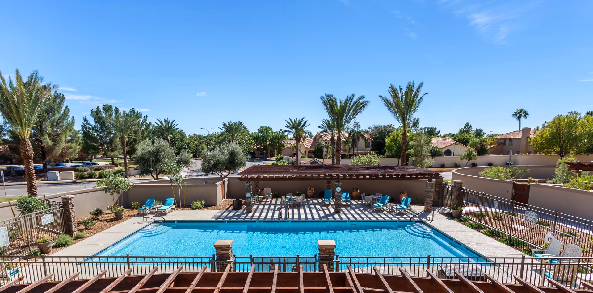 Swimming pool with lounge chairs and a shaded pergola surrounded by palm trees and fencing under a clear blue sky.
