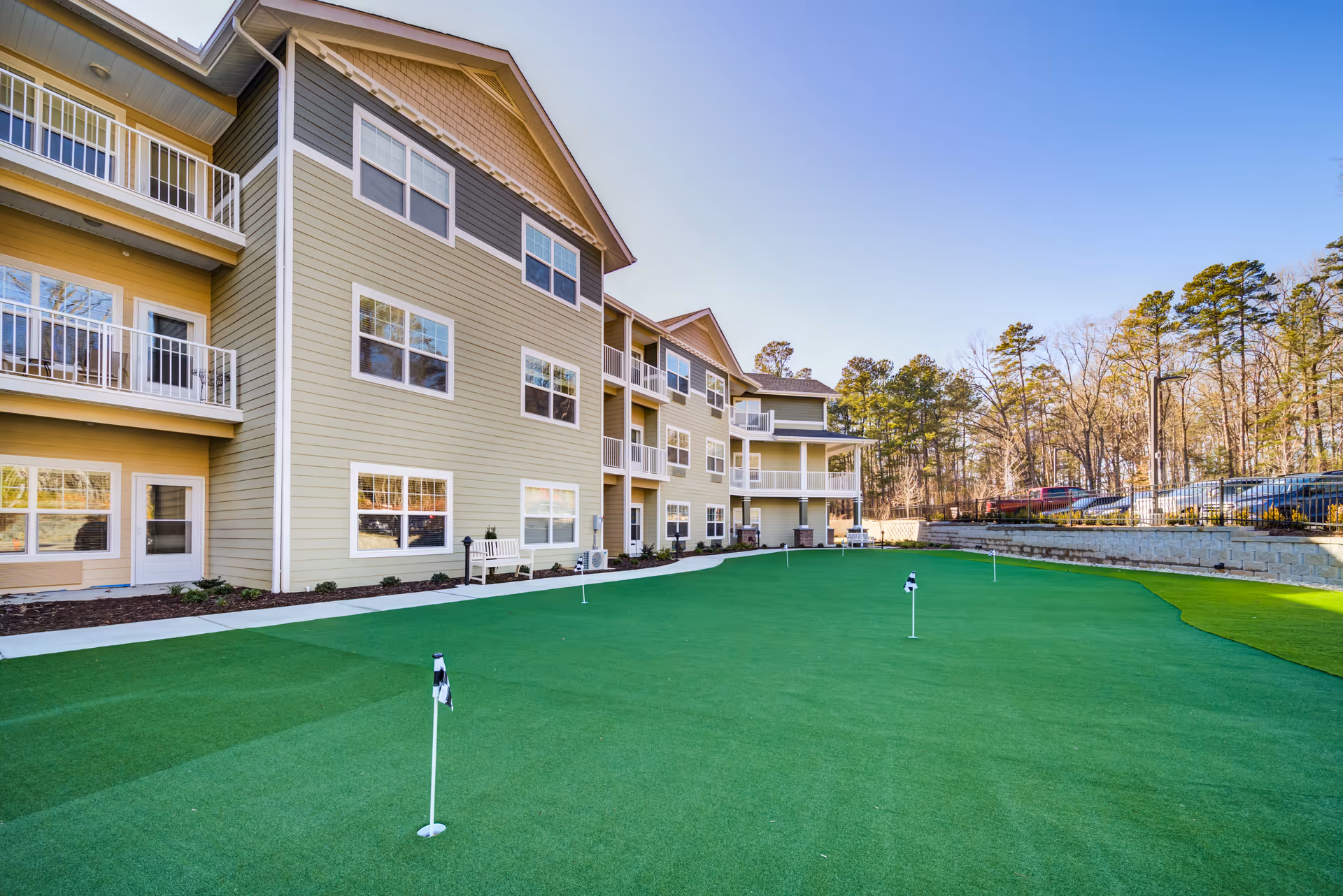 Exterior view of a multi-story residential building with balconies overlooking a well-maintained putting green with several golf holes and flags. Trees and a parking area are visible in the background under a clear blue sky.