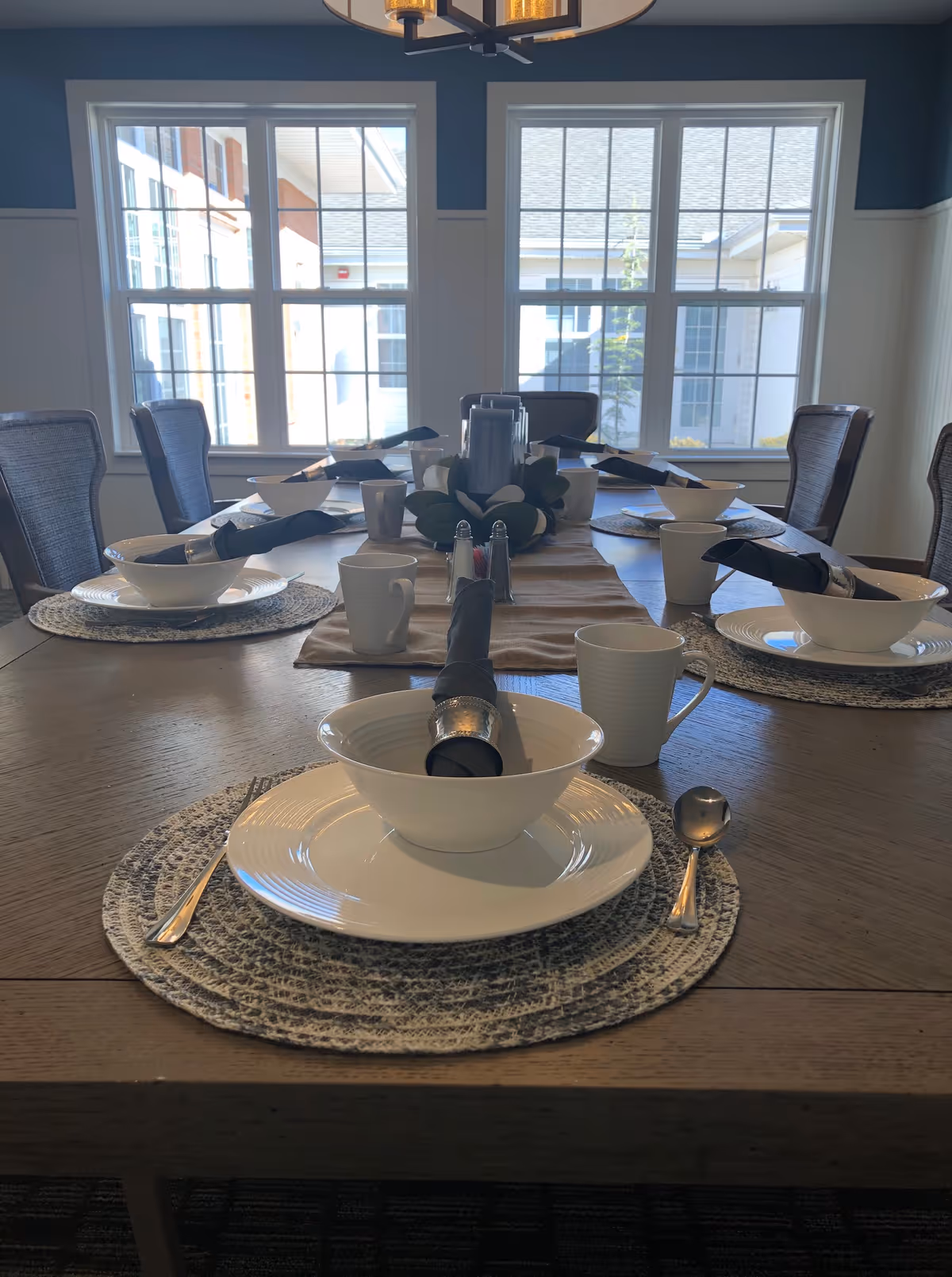 A dining table set for six with white plates, bowls, cups, and silverware. Each place setting has a black napkin with a silver napkin ring. The table has a beige runner and a centerpiece with candles and greenery. Large windows in the background let in natural light.