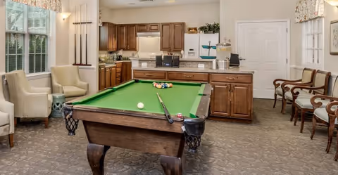 Interior room featuring a pool table with a cue stick and balls set up for a game. The room has wooden cabinetry along the back wall with a countertop, a water dispenser, and some decorative plants. There are several chairs arranged along the walls, two armchairs near a window with blinds, and a rack holding pool cues. The space is well-lit with natural light coming through the window and wall-mounted lights.