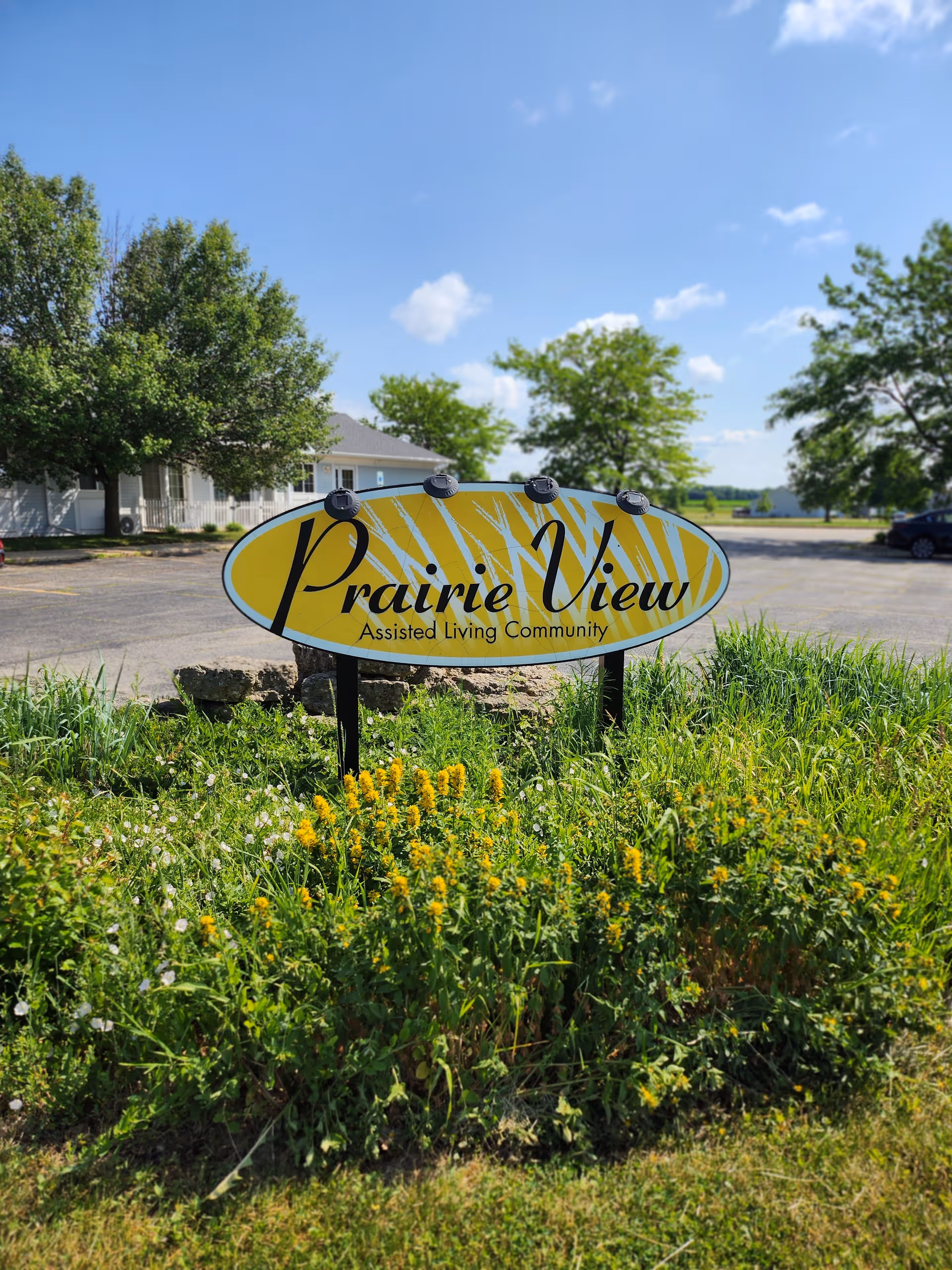 Oval yellow sign reading "Prairie View Assisted Living Community" in a grassy planting area with a parking lot, building, and trees behind it.