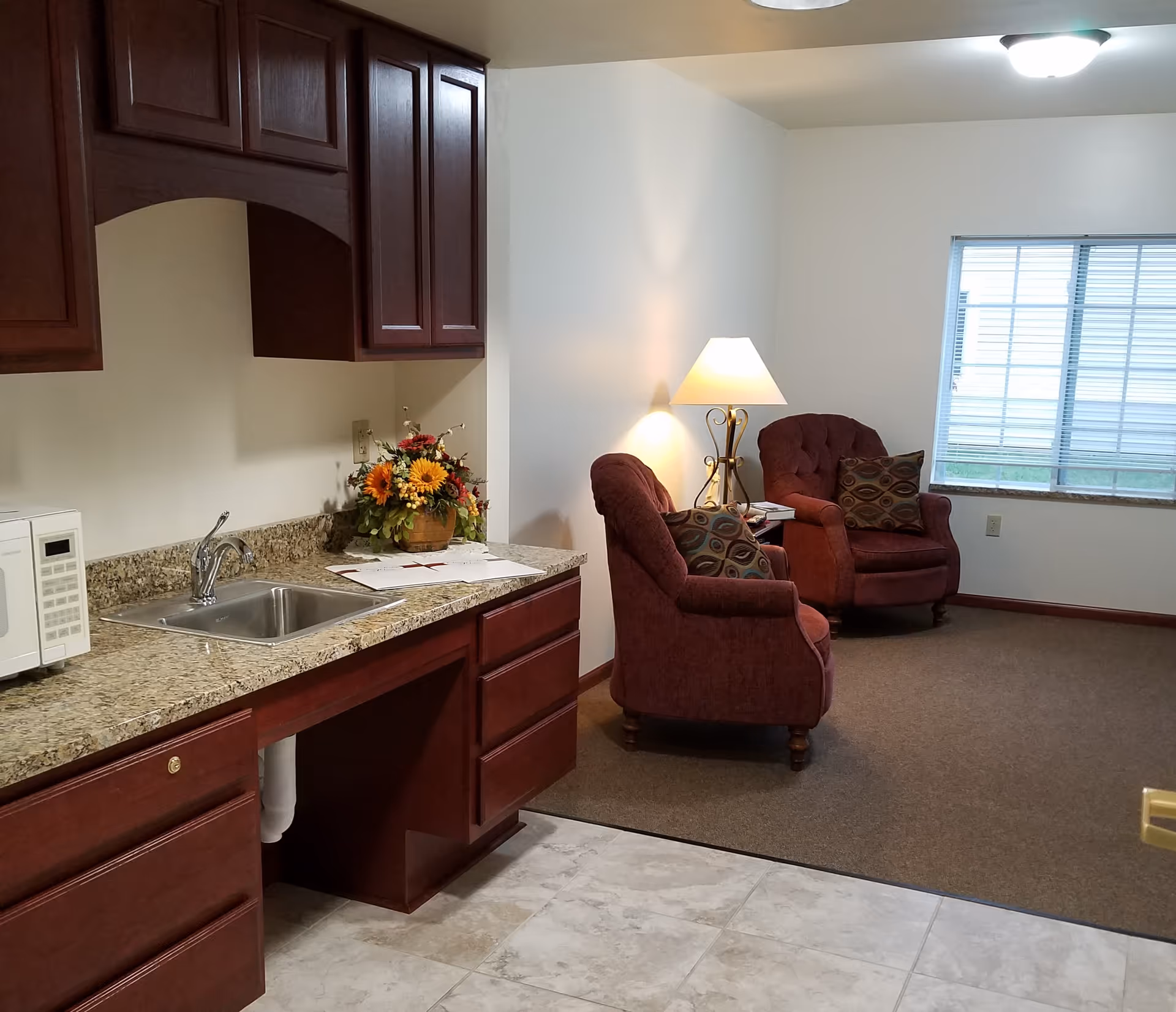 Interior view of a senior living facility room showing a kitchenette area with a sink, microwave, granite countertop, and wooden cabinets. Adjacent to the kitchenette is a small sitting area with two upholstered armchairs, a side table with a lamp, and a window with blinds letting in natural light.