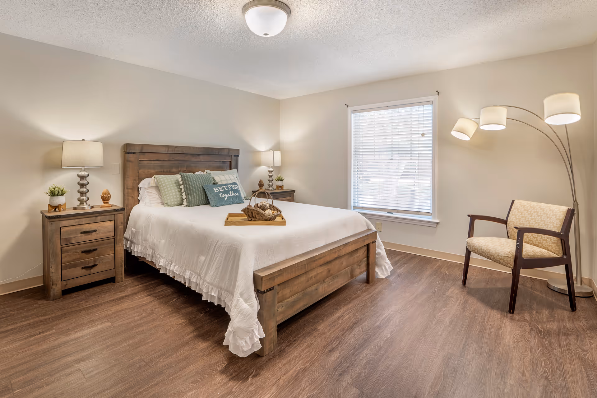 A cozy bedroom with a wooden bed frame and white bedding, decorated with green and white pillows including one that says 'BETTER together'. There are two matching wooden nightstands with lamps and small plants on each side of the bed. A window with blinds is on the right wall, next to a beige upholstered chair and a modern floor lamp with four lights. The floor is wood, and the walls are painted light beige.