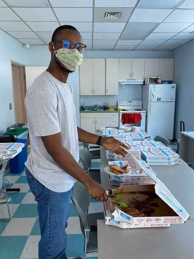 A man wearing a face mask and glasses is serving himself a slice of pizza from an open pizza box on a table in a kitchen or break room. Several closed pizza boxes are stacked on the table. The room has a refrigerator, stove, sink, and cabinets in the background.