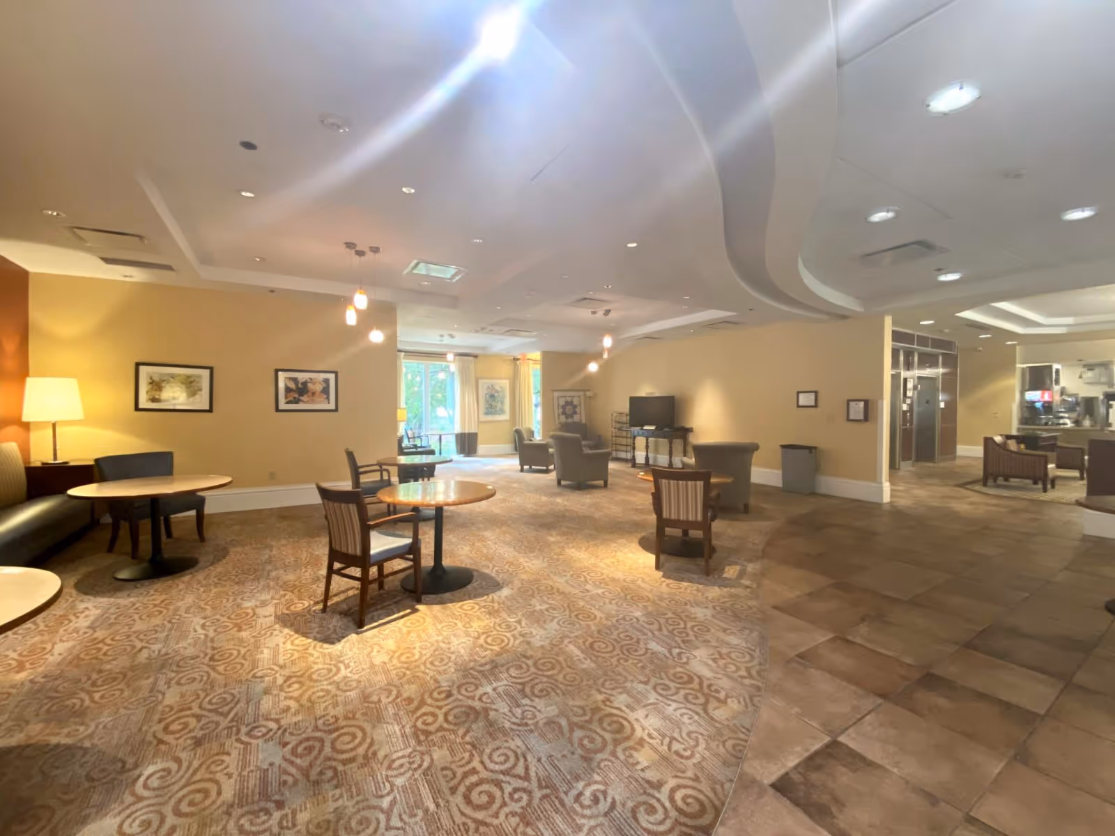Spacious communal lounge with round tables and chairs, armchairs, a television and warm ambient lighting.