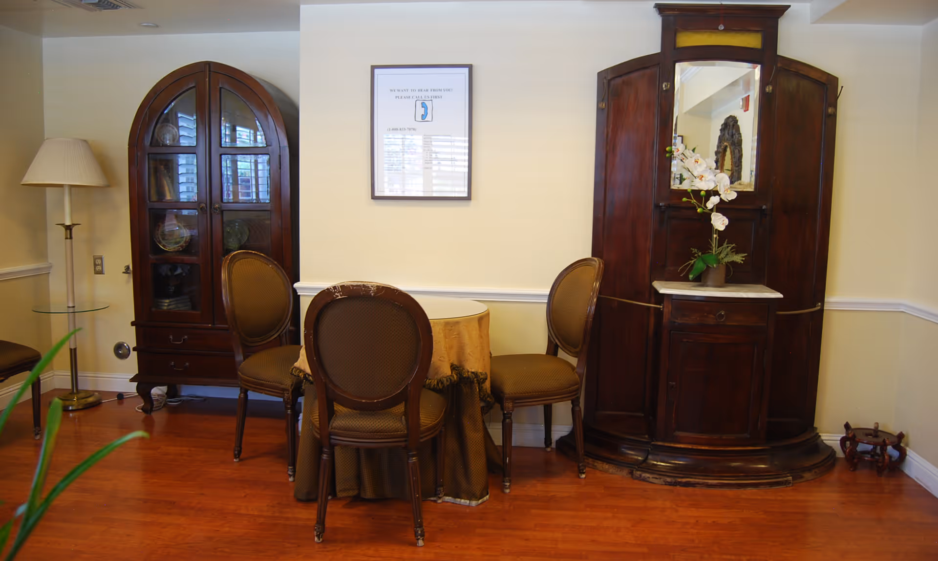 A cozy interior corner of a senior living facility with a small round table covered with a brown tablecloth and surrounded by three brown upholstered chairs. To the left is a tall wooden cabinet with glass doors displaying decorative plates, and a floor lamp with a beige shade. On the right is a large wooden cabinet with a mirror and a vase with white flowers on top. The floor is wooden, and the walls are painted light yellow with white trim. A framed sign is hung on the wall above the table.