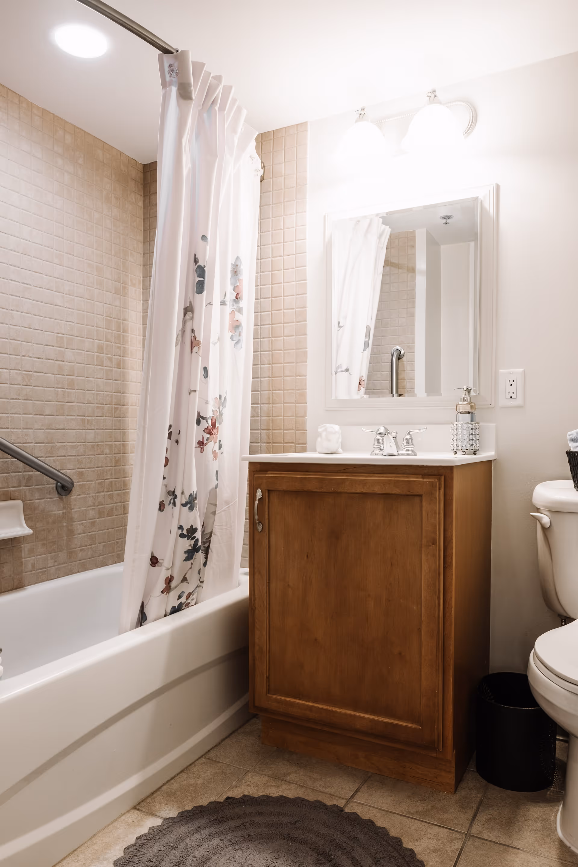 A clean bathroom featuring a bathtub with a floral shower curtain, beige tiled walls, a wooden vanity with a white countertop and sink, a mirror above the sink, a toilet, and a round gray bath mat on the tiled floor.