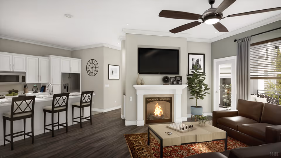 A modern open-concept living room and kitchen area in a senior living facility. The living room features a brown leather sofa, a light wood coffee table with a chessboard and glasses on it, a fireplace with a fire burning, and a flat-screen TV mounted above the fireplace. There is a large potted plant next to the fireplace and a window with blinds and curtains letting in natural light. The kitchen has white cabinets, a stainless steel refrigerator, a microwave, and a countertop with three high chairs. The floor is dark wood, and the walls are painted in neutral tones. A ceiling fan with lights is mounted on the ceiling.