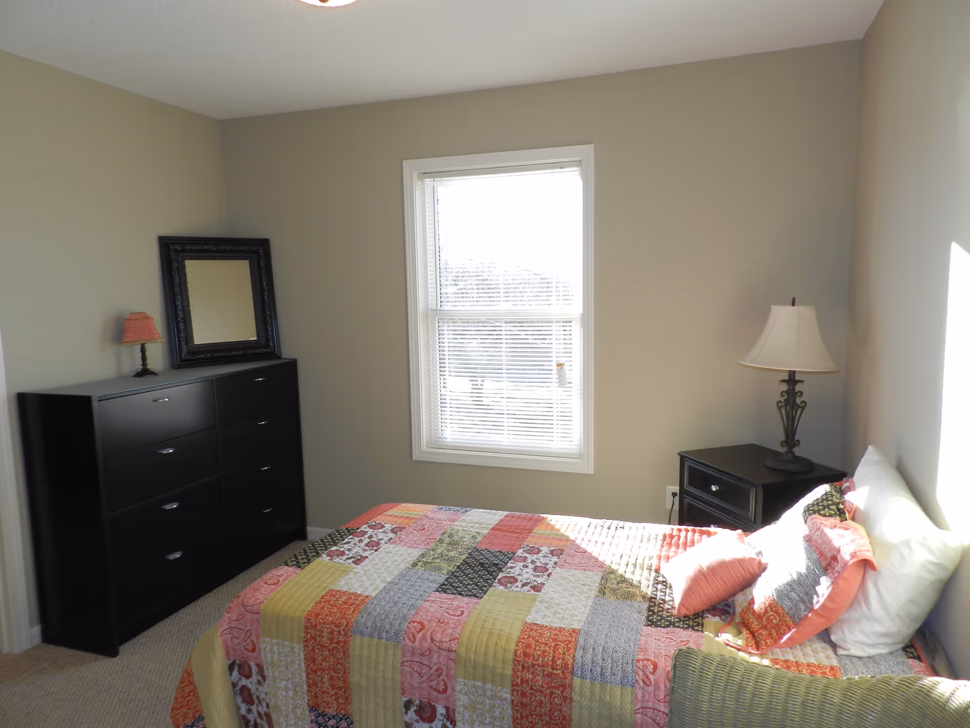 Sunlit bedroom with a twin bed covered in a colorful patchwork quilt, a dresser with a framed mirror, and a nightstand with a lamp.