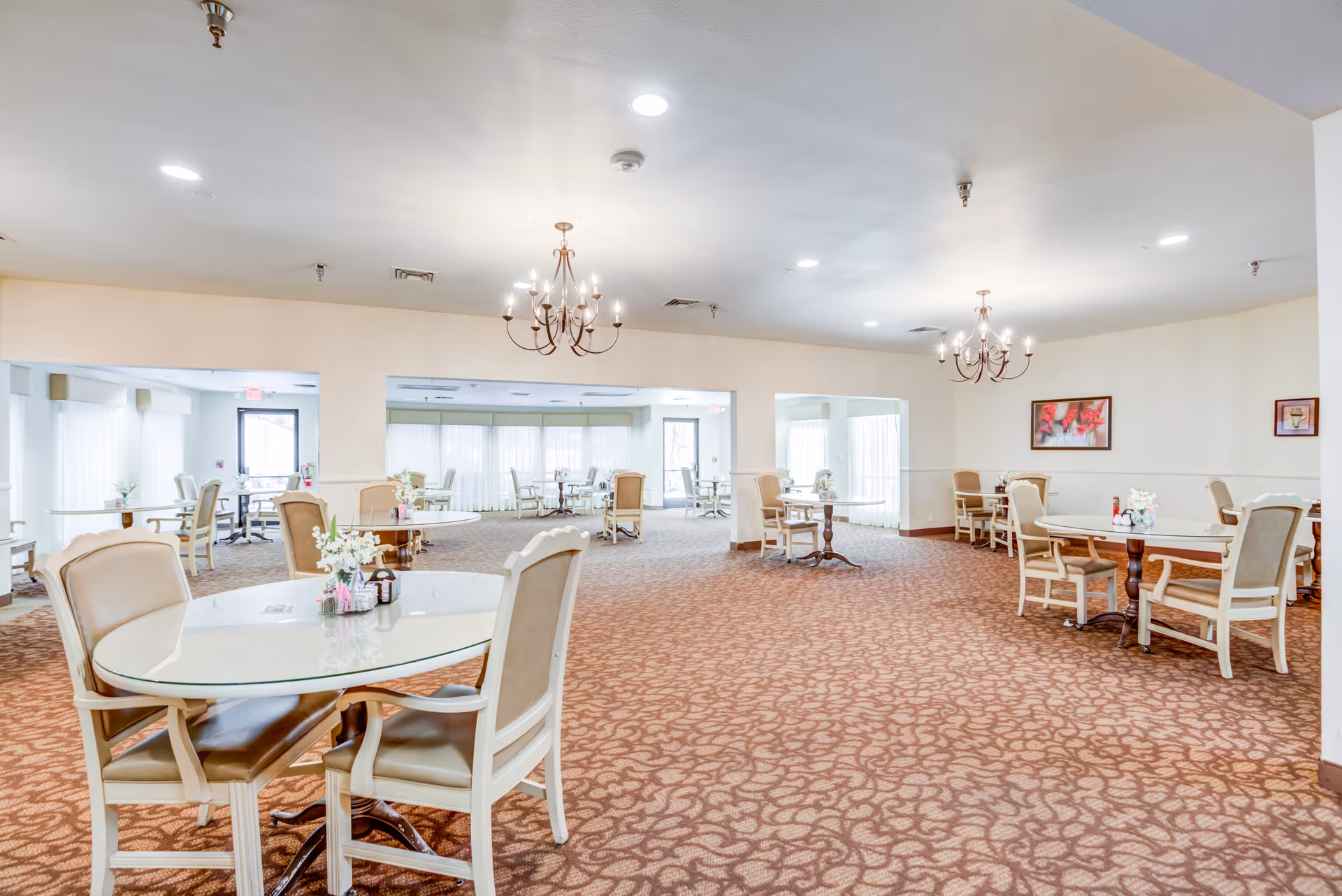Spacious senior living dining area with multiple round tables and chairs, chandeliers, and patterned carpet.
