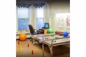 Rehabilitation room with parallel bars, colorful therapy balls and cones, an office chair with a laptop, and a medical chart by the windows.