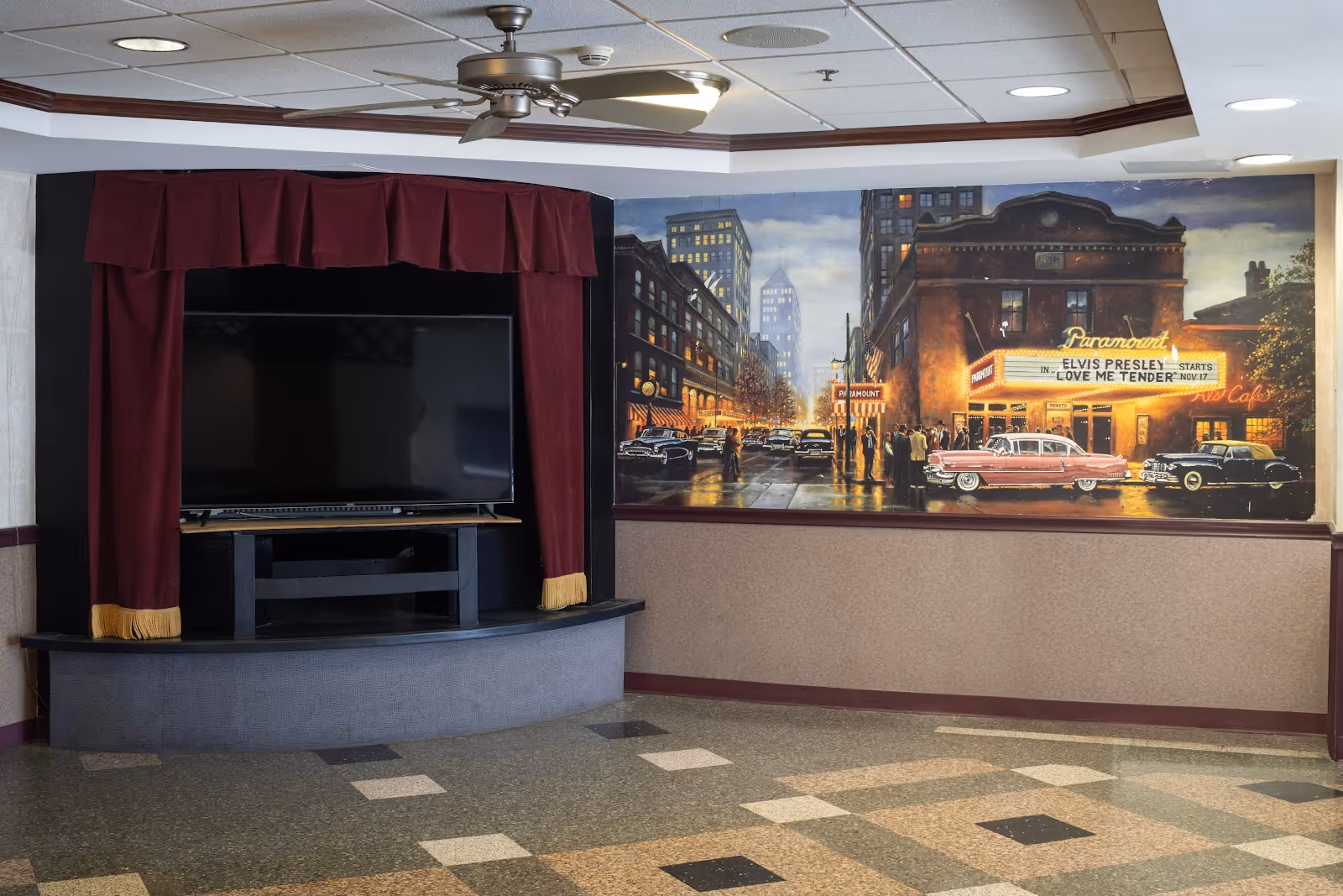 An interior common area with a large flat-screen TV set into a black entertainment center framed by red curtains with gold tassels. To the right, there is a mural depicting a vintage city street scene at night with classic cars and a theater marquee advertising Elvis Presley's 'Love Me Tender'. The floor has a patterned tile design, and the ceiling has recessed lighting and a ceiling fan.