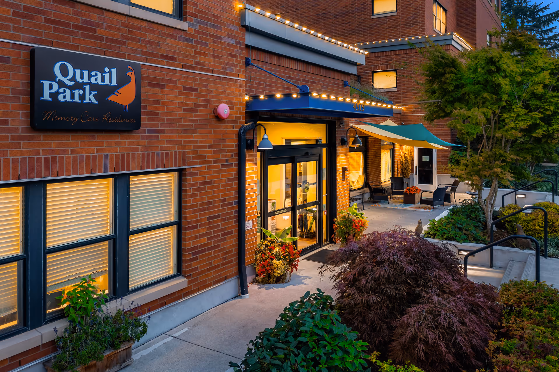 Entrance to the Quail Park memory care residence with a lit awning, brick façade, glass doors, and landscaped walkway.