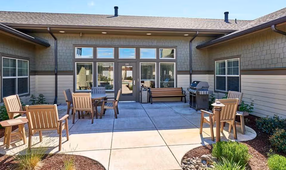 Outdoor patio area at Waterhouse Ridge Memory Care with wooden chairs and tables arranged on a concrete surface, a bench, a barbecue grill, and surrounding landscaping with plants and mulch beds.