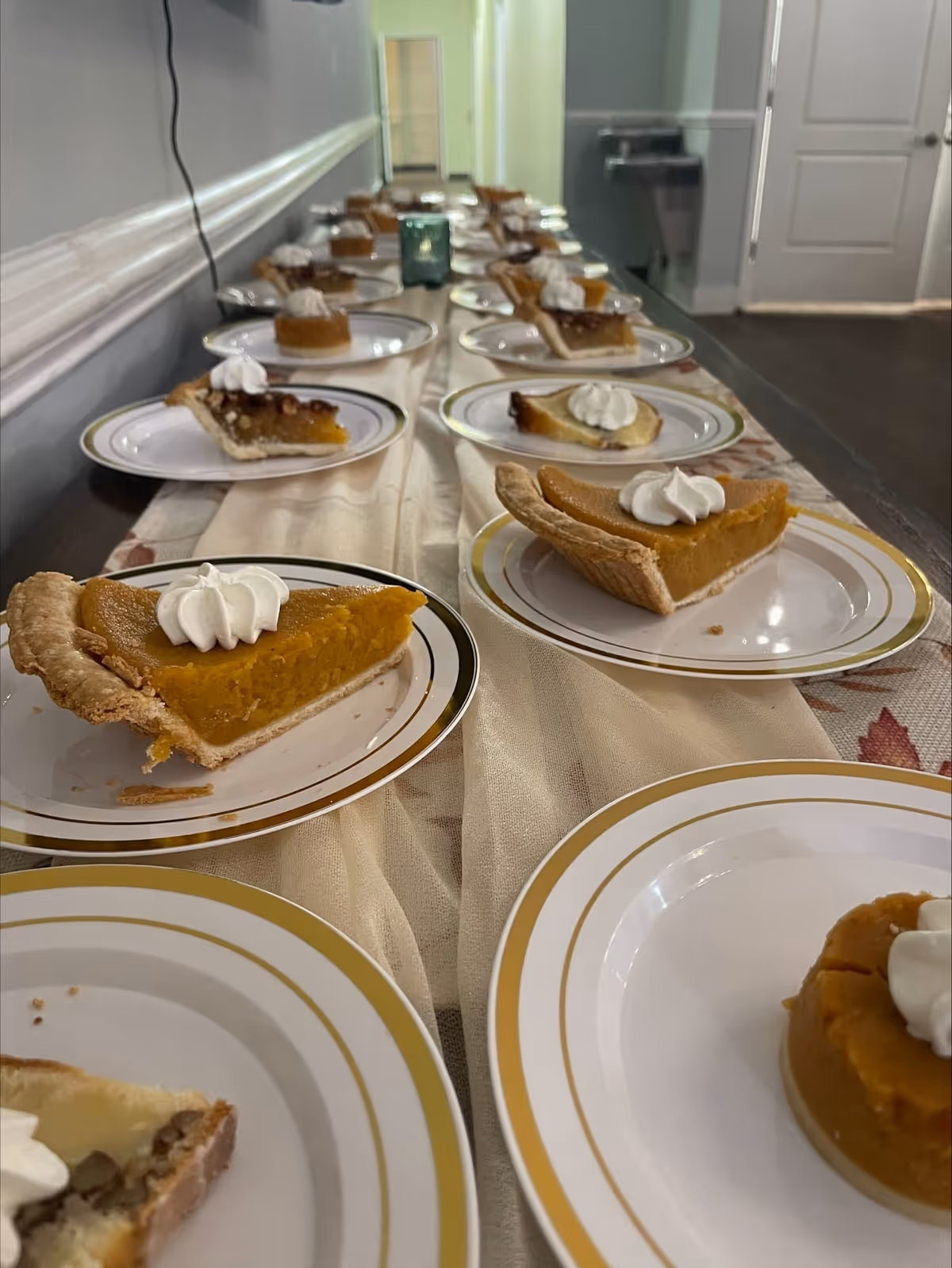 A long table covered with a beige tablecloth displaying multiple plates of pie slices topped with whipped cream, arranged in a row in a hallway or dining area with a water fountain and closed doors in the background.
