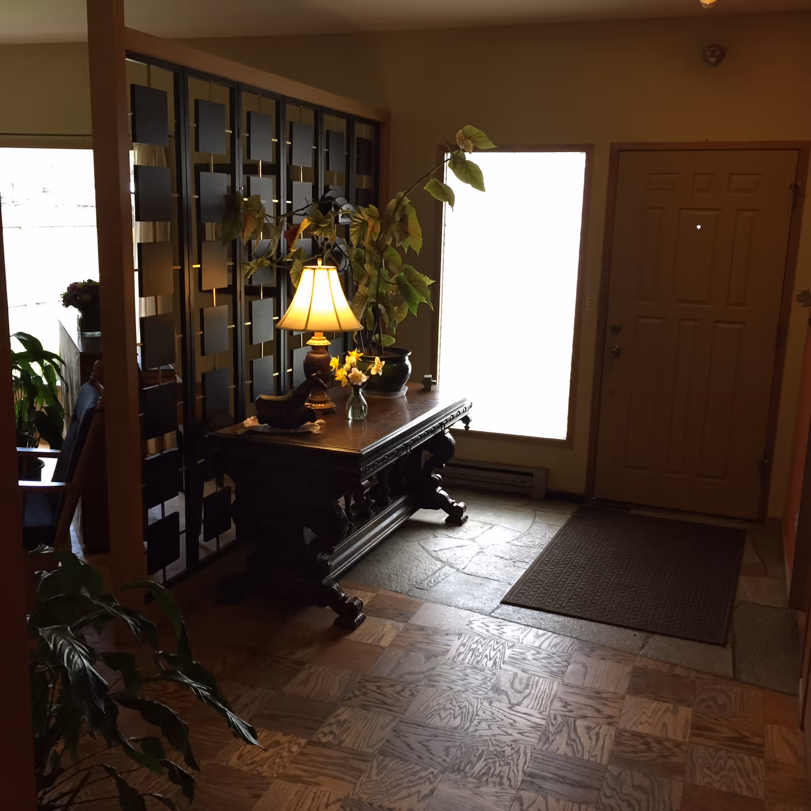 A warmly lit entry foyer with an ornate wooden console table holding a lamp and plants, a decorative room divider, and a closed front door.