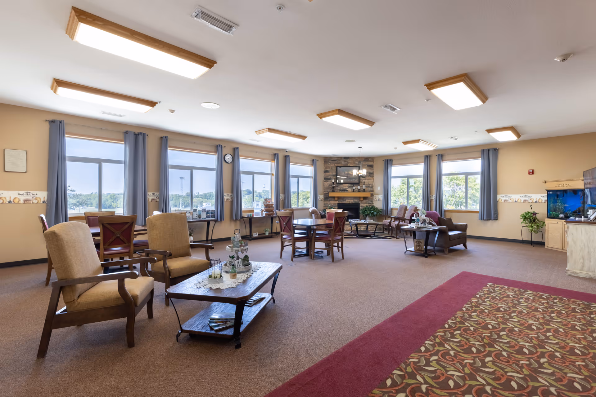 Spacious senior living common area with multiple seating arrangements including armchairs and tables. Large windows with blue curtains allow natural light to fill the room. A stone fireplace is centered on the far wall, flanked by additional seating. The room has beige walls with a decorative border and a patterned carpet near the entrance. A large fish tank is visible on the right side.