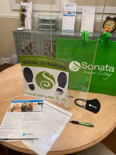 A round wooden table with a clear plastic divider on it. On the table are a green Sonata Senior Living tote bag, a social distancing sign asking to stay 6 feet apart, two small hand sanitizer bottles, a black face mask with the Sonata logo, a green and black pen, and some informational brochures and papers. In the background, there is a cabinet with glass doors and some standing brochures on top.