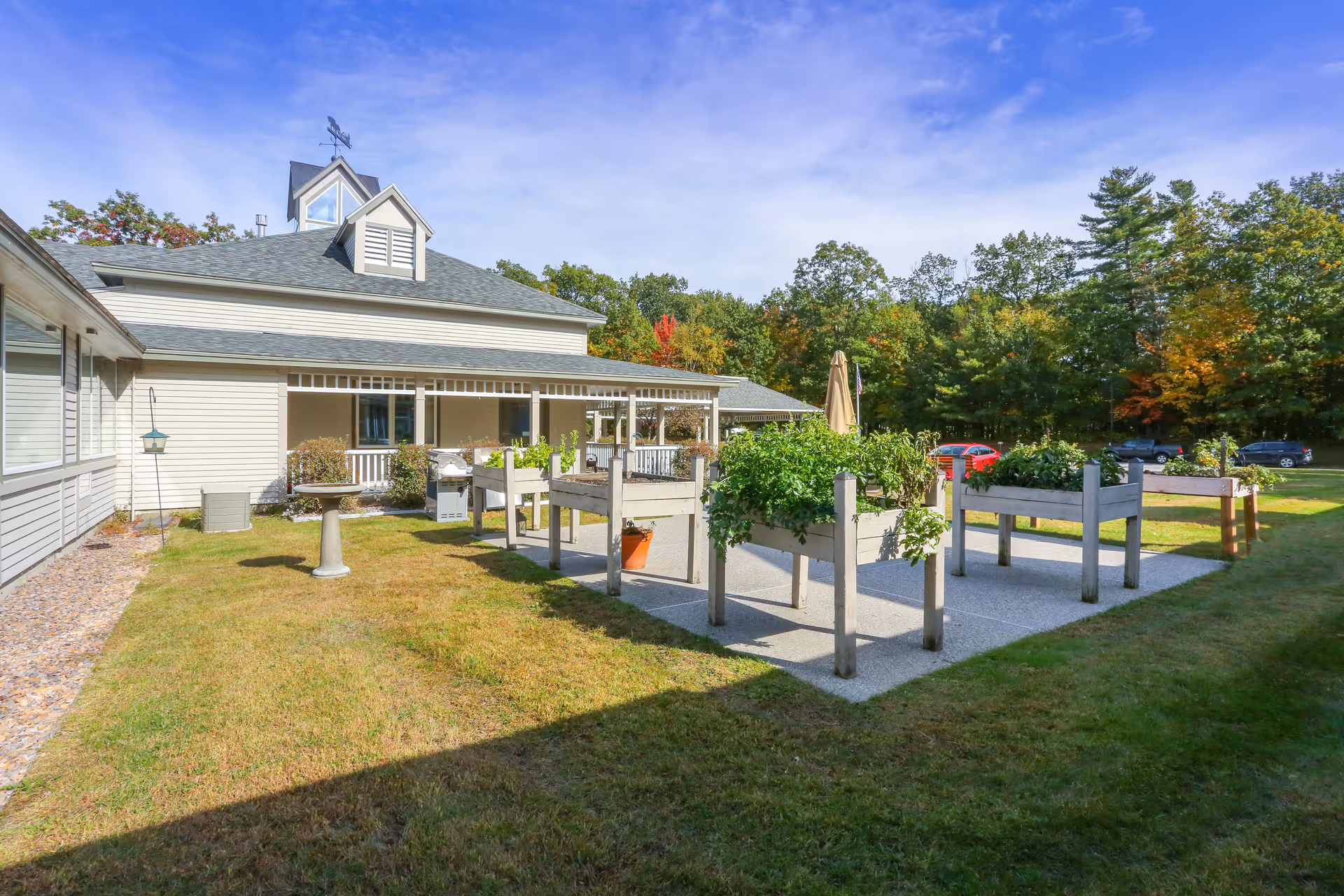 Outdoor garden area at Elderwood at Burlington featuring raised garden beds with green plants, a birdbath, and a building with a covered porch in the background under a blue sky.