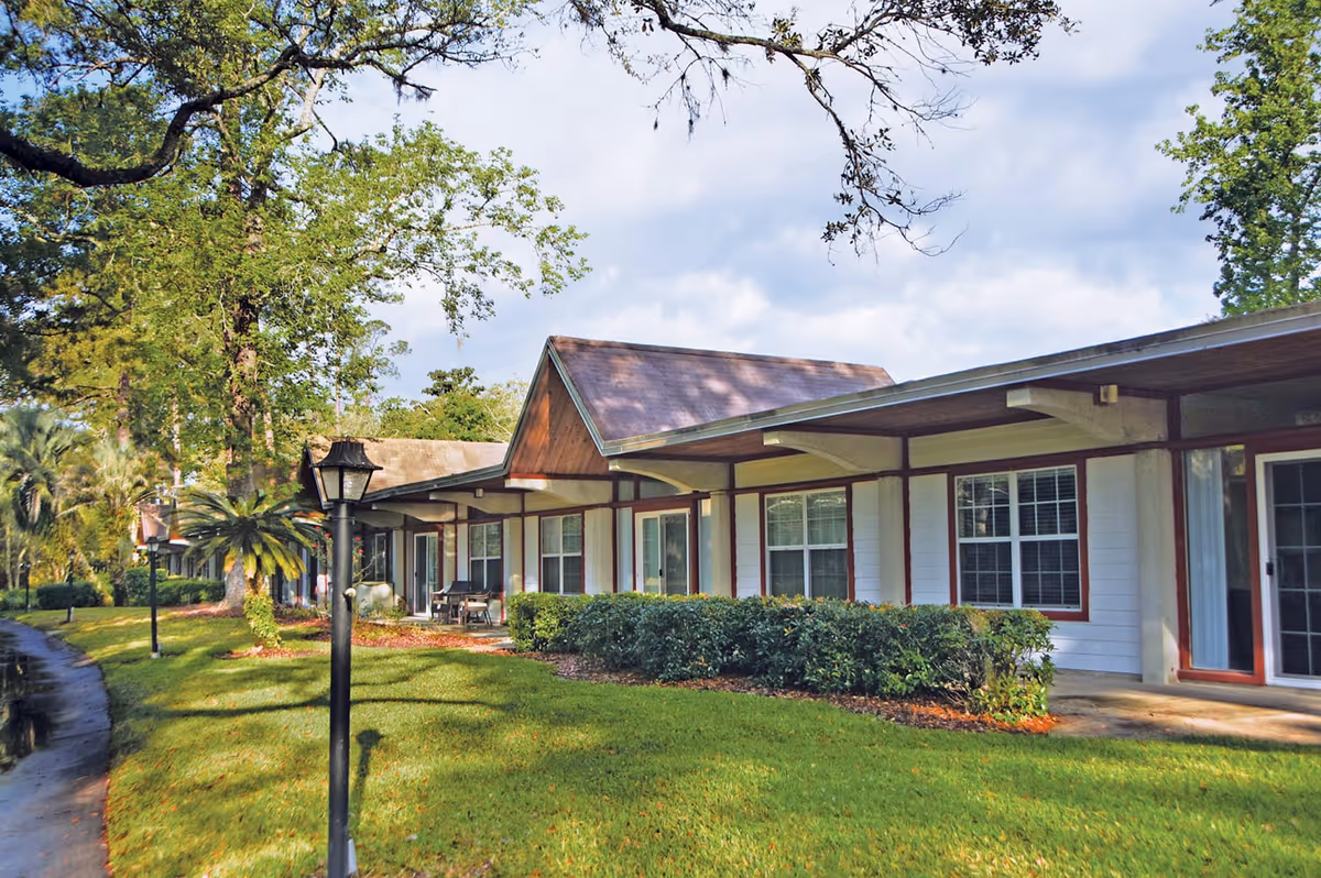 Exterior view of a single-story building at Westminster Woods on Julington Creek with a sloped roof, multiple windows, and a covered walkway. The building is surrounded by green grass, bushes, tall trees, and a paved pathway with black lamp posts.