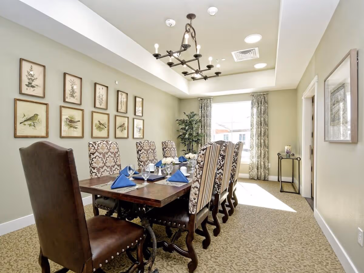 Sunlit dining room with a long wooden table set with blue napkins and patterned high-back chairs under a chandelier.