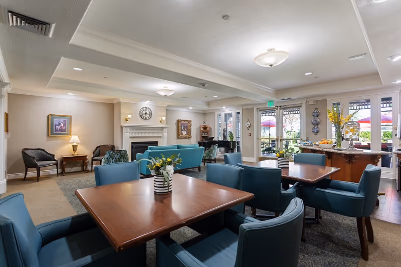 A spacious and well-lit common area in a senior living facility featuring multiple wooden tables with blue upholstered chairs. The room includes a fireplace with a clock above it, framed artwork on the walls, and a side table with a lamp. Large windows and glass doors open to an outdoor patio with red umbrellas. There is also a counter with a vase of yellow flowers and a fruit bowl.