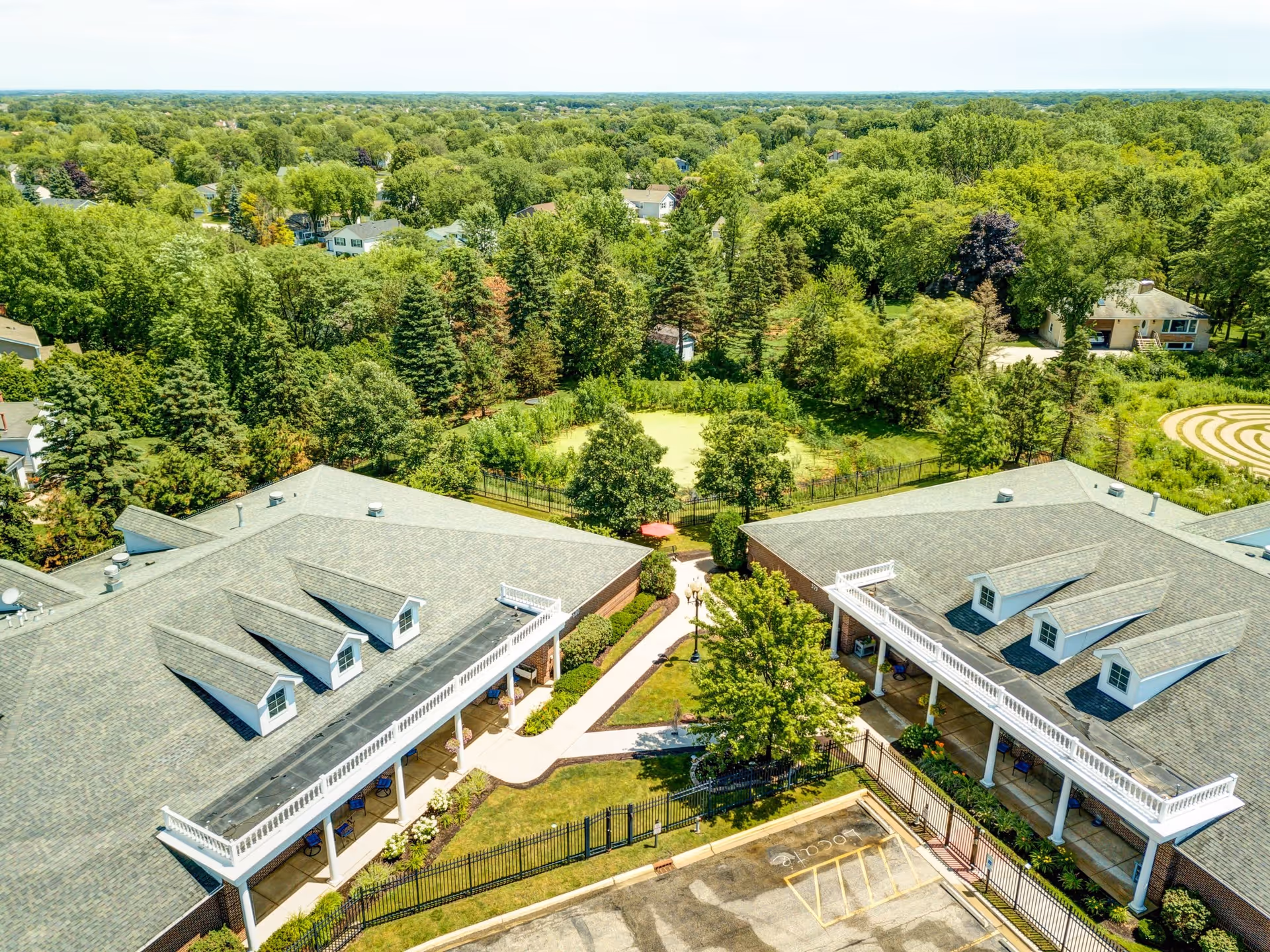 Aerial view of The Auberge at Lake Zurich senior living facility showing two large buildings with gray roofs and white trim, connected by a walkway surrounded by green lawns and trees. The background features a dense forested area and residential houses.