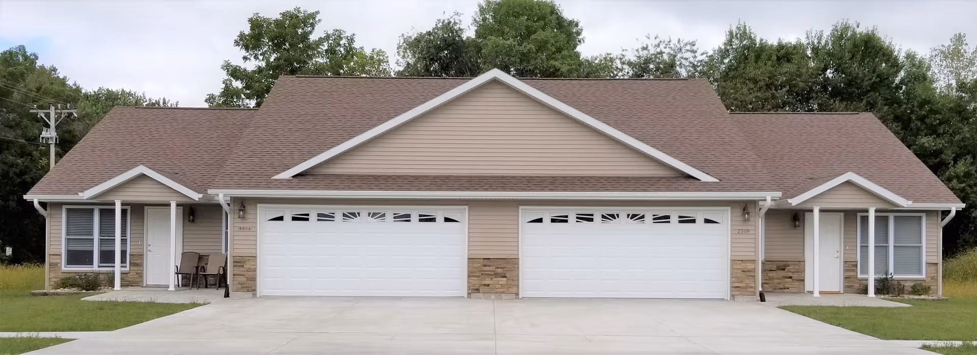 Exterior view of a single-story duplex building with beige siding, brown shingled roof, two white garage doors, and small covered porches with chairs on each side. The building is surrounded by green trees and has a concrete driveway in front.
