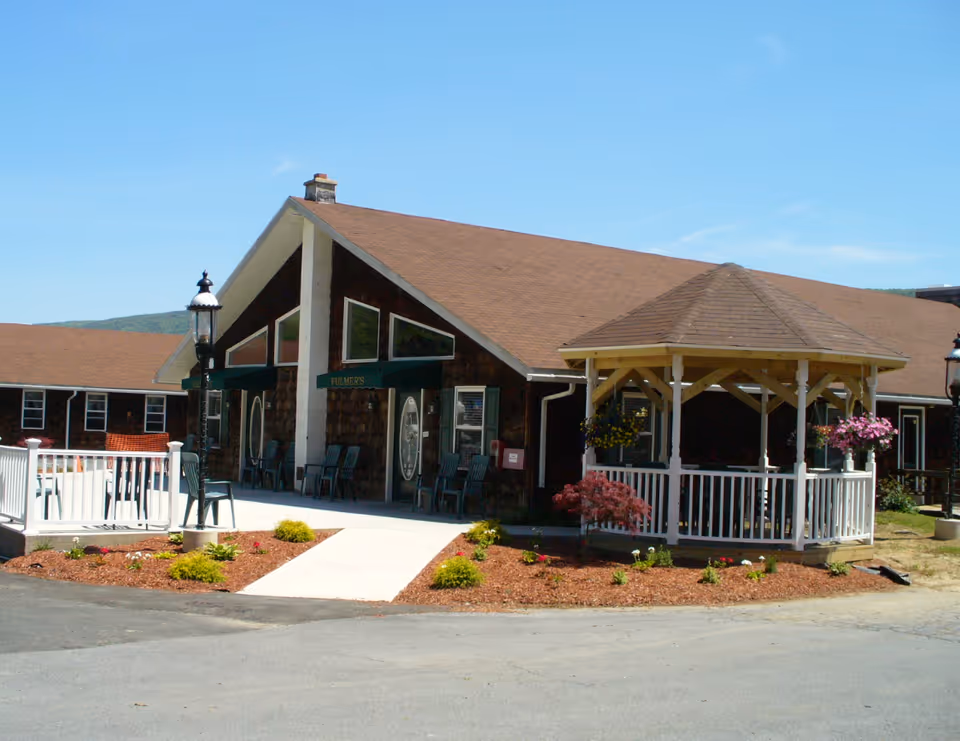 Exterior view of Fulmer's Personal Care Home, showing a brown building with a sloped roof, a white gazebo with hanging flower baskets, a paved walkway, and landscaped garden beds with small shrubs and flowers under a clear blue sky.