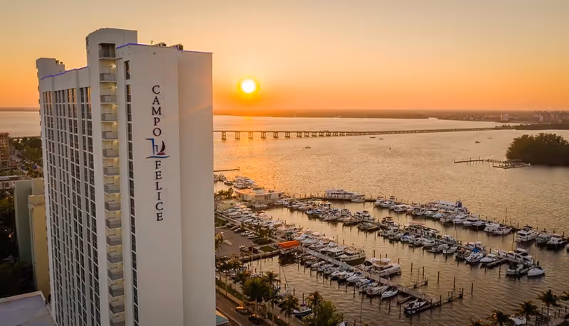 Tall building with the name Campo Felice on the side, overlooking a marina filled with boats at sunset with a bridge and water in the background.