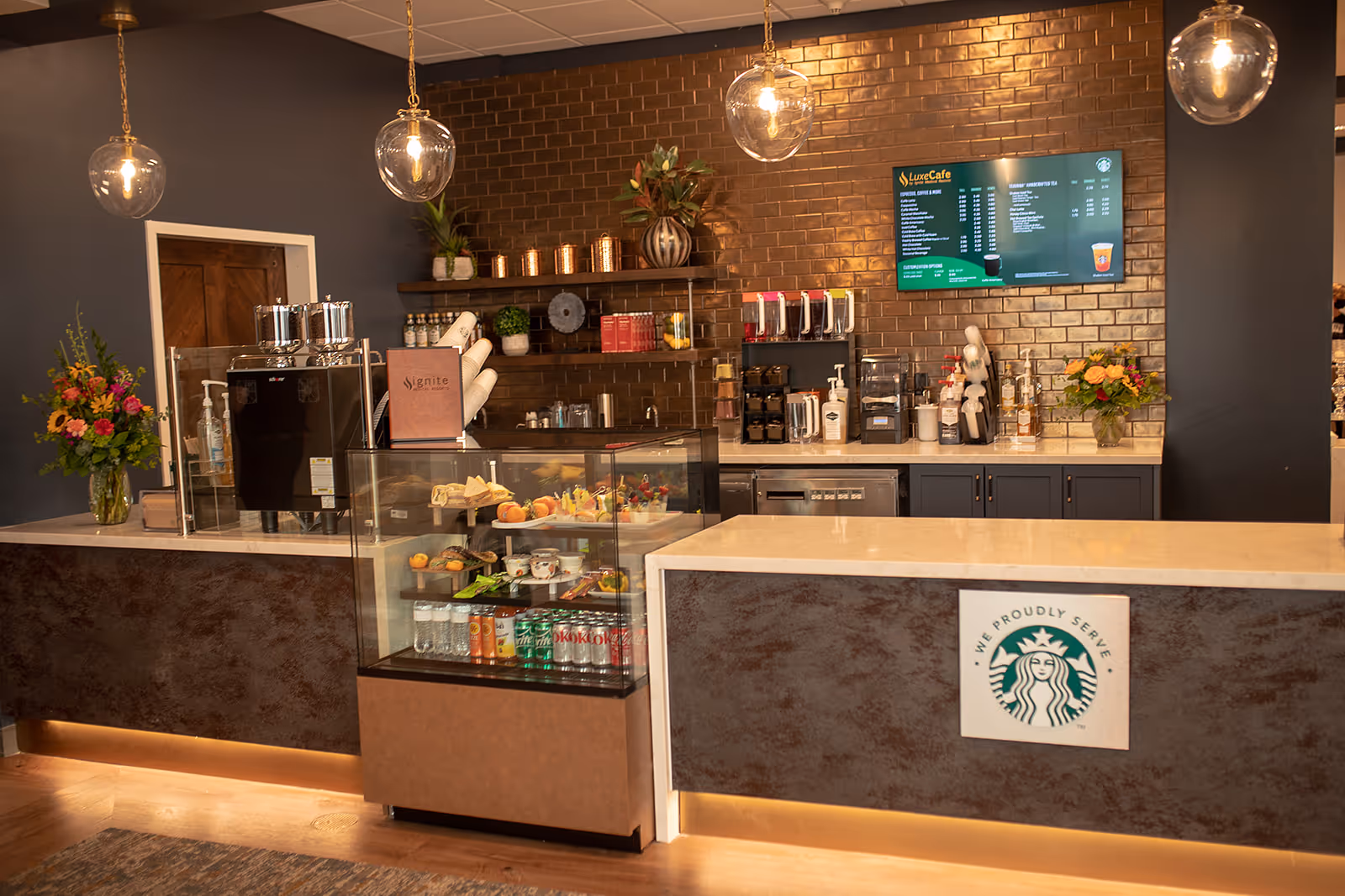 Interior view of a modern cafe counter area with a glass display case containing snacks and beverages. Behind the counter are coffee machines, dispensers, shelves with decorative items, and a digital menu board on a brown tiled wall. There are hanging pendant lights above the counter and flower arrangements on both sides.