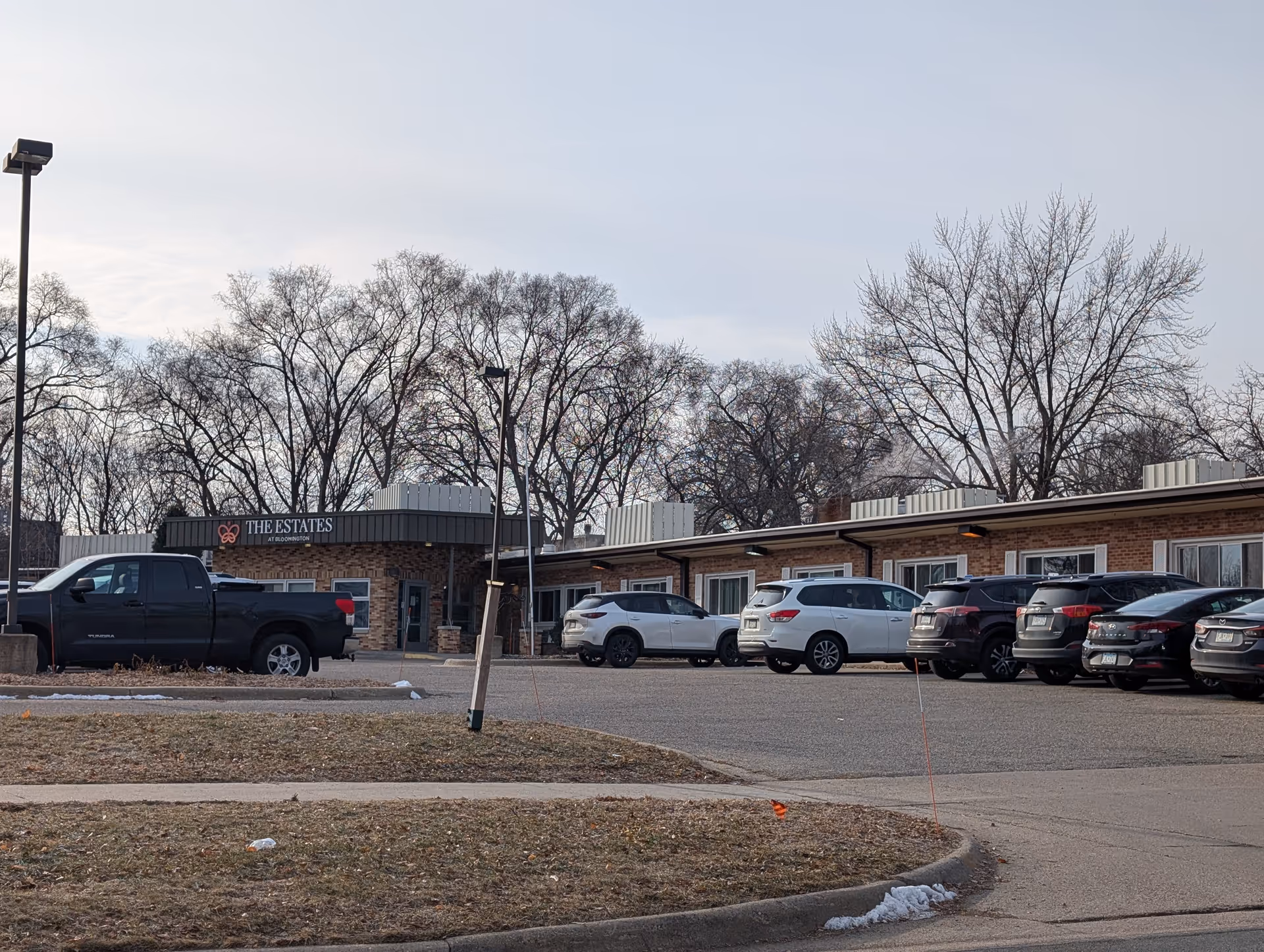 Exterior view of The Estates at Bloomington senior living facility showing a single-story brick building with several parked cars in front and leafless trees in the background under a cloudy sky.