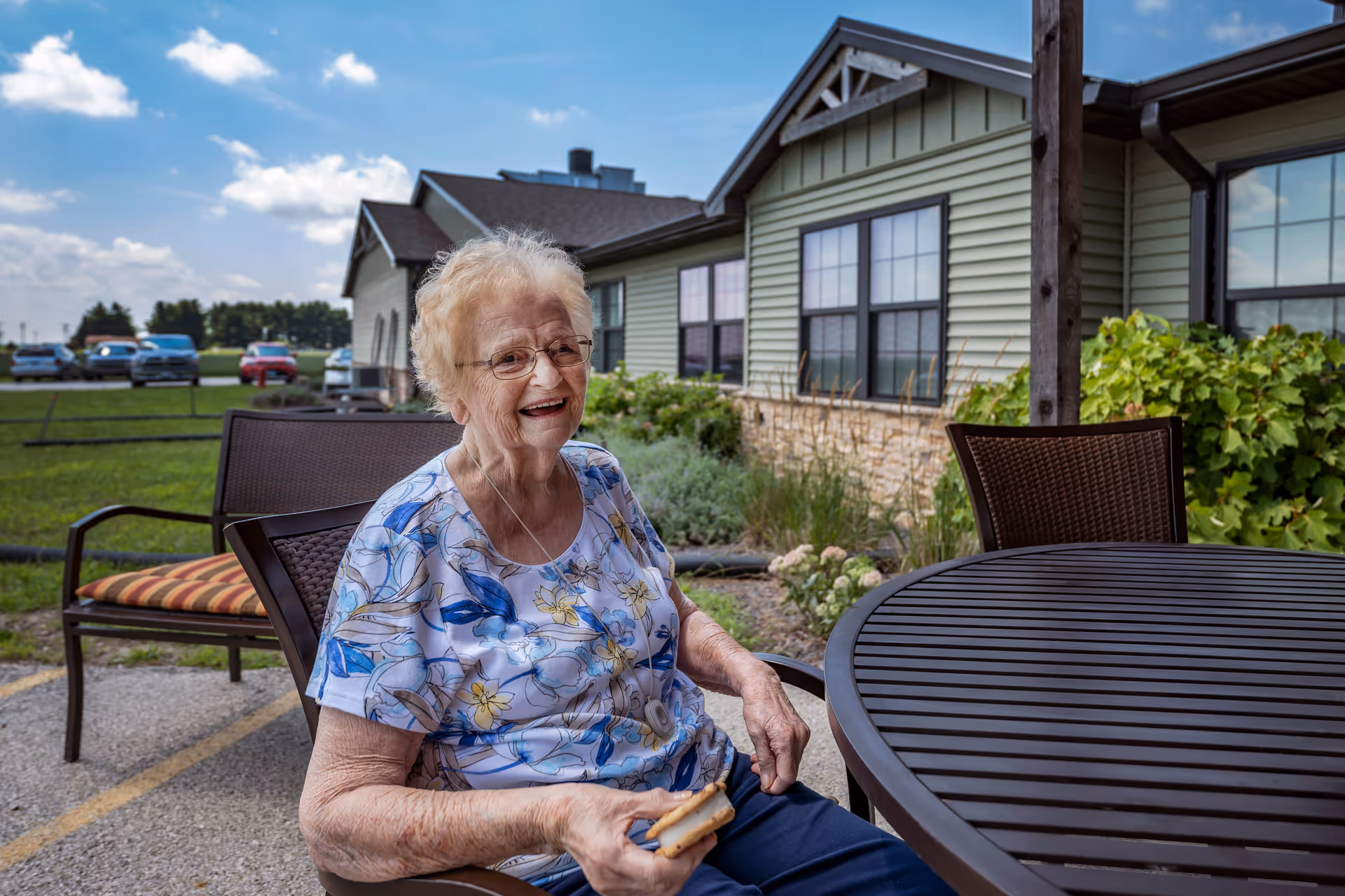 An elderly woman with glasses and a floral shirt is sitting outdoors at a round metal table, smiling and holding a sandwich. Behind her is a building with green siding and multiple windows, some greenery, and a parking area with several cars under a partly cloudy sky.