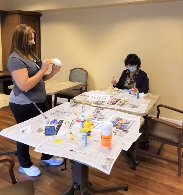 Two women engaged in a painting activity at a senior living facility. One woman is standing and painting a white object, while the other woman, wearing a face mask, is seated and painting a small item. The tables are covered with newspapers and various painting supplies including brushes, paint bottles, and a disposable coffee cup. The room has wooden flooring and simple furniture.