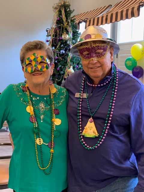Two elderly individuals wearing colorful Mardi Gras masks and bead necklaces stand together indoors in front of a decorated Christmas tree and window with striped valance. One person is wearing a green top and the other a purple top with a white hat.