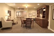 A cozy interior room with beige walls and carpeted floor featuring a wooden dining table with four chairs in the center. To the left, there are two cushioned armchairs with a small side table and lamp between them. On the right side, there is a large wooden built-in cabinet with shelves and framed pictures on the wall above it. The room is softly lit with ceiling lights and has a window with blinds in the background.