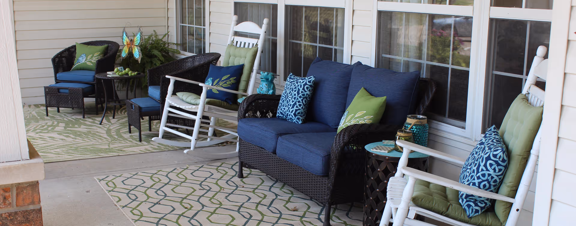 Outdoor patio area with wicker furniture including a loveseat and chairs with blue and green cushions, white rocking chairs, small tables, decorative pillows, and two patterned outdoor rugs on a concrete floor next to a building with large windows.