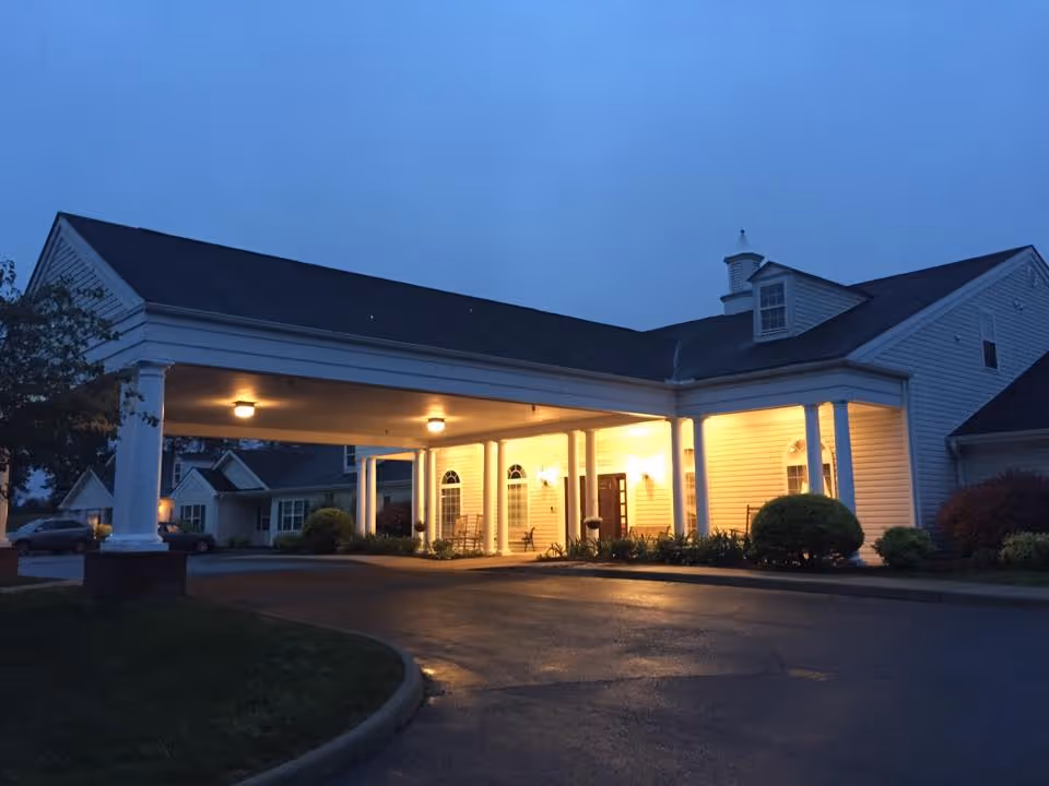 Exterior view of a senior living facility entrance at dusk with a covered driveway lit by ceiling lights. The building is white with columns and has a well-maintained garden area near the entrance.