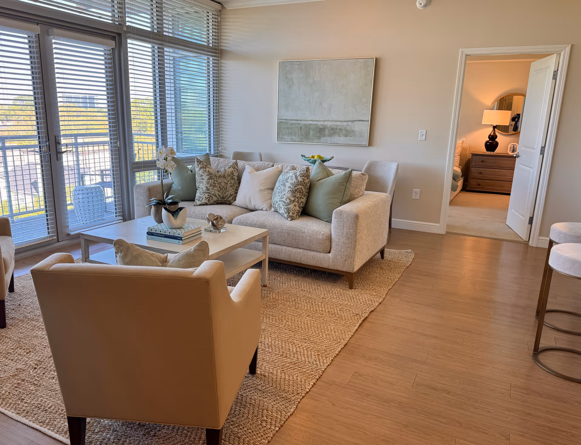 A bright living room with large windows covered by blinds, a beige sofa with multiple decorative pillows, a beige armchair, a light-colored coffee table with books and a plant, a textured area rug, and a doorway leading to a bedroom with a lamp and dresser visible.