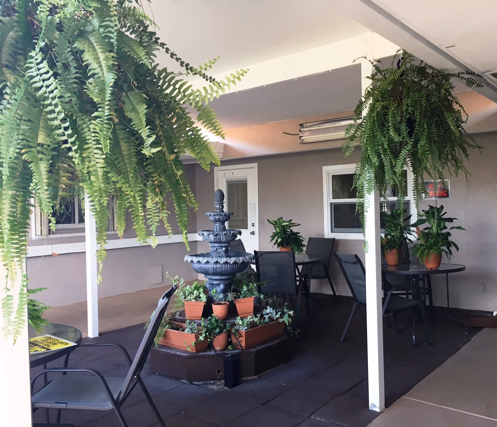 Covered outdoor patio area with hanging ferns, a tiered water fountain surrounded by potted plants, several tables and chairs, and a 'No Smoking' sign on the wall.