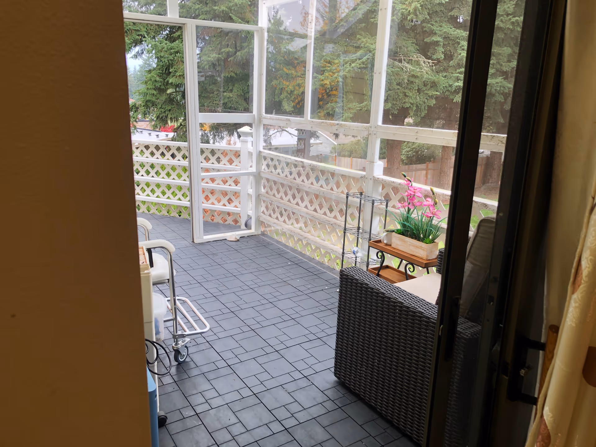 View of a screened-in porch with black tiled flooring, a wicker chair with cushions, a small table holding a planter with pink flowers, and a metal rack. The porch has white lattice railing and is surrounded by tall green trees outside.