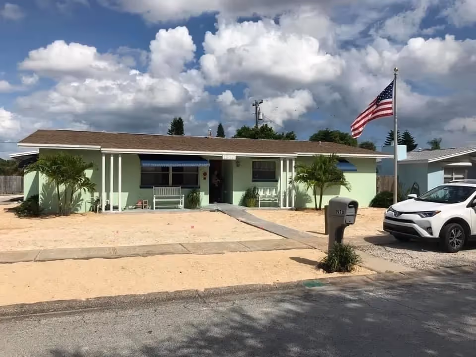 Single-story light green house with a front porch, entry ramp, mailbox, American flag, and a parked white SUV under a cloudy sky.