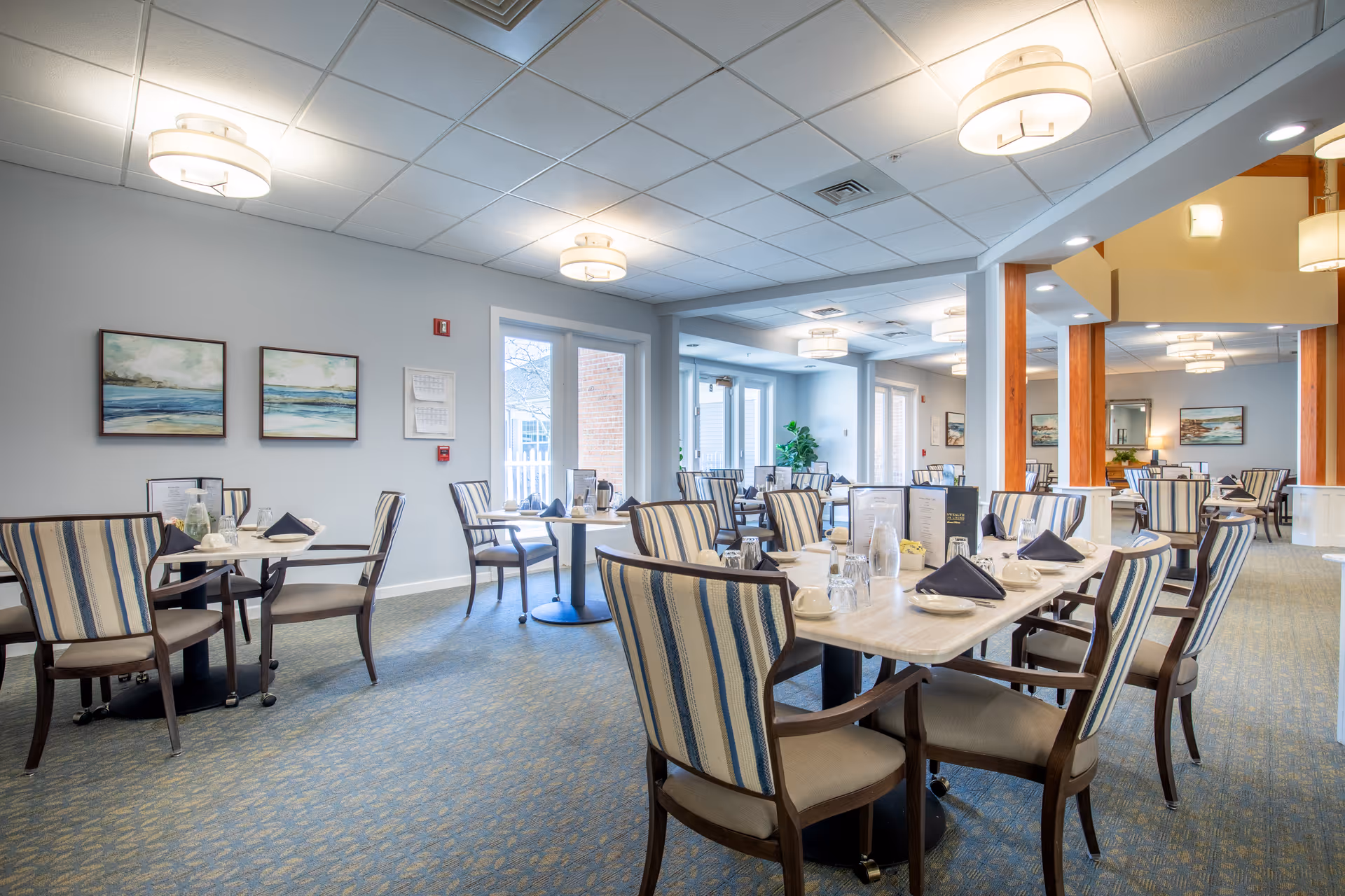A bright and spacious dining room in a senior living facility with multiple tables set with plates, glasses, napkins, and menus. The room features striped upholstered chairs, soft blue walls, carpeted floors, and modern ceiling lights. Large windows and glass doors allow natural light to enter, and framed artwork decorates the walls.