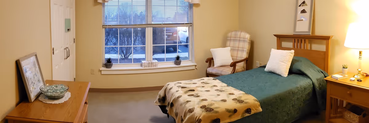 A cozy senior living bedroom with a single bed covered in a green bedspread and a beige blanket with pinecone patterns. There is a wooden headboard, a white pillow, and a wooden nightstand with a lamp, a small plant, and some books. A cushioned armchair with a plaid pattern and a white pillow is placed near a window with blinds. A wooden dresser with a glass bowl and a framed picture is on the left side of the room. The walls are painted light yellow, and the room has a calm and inviting atmosphere.