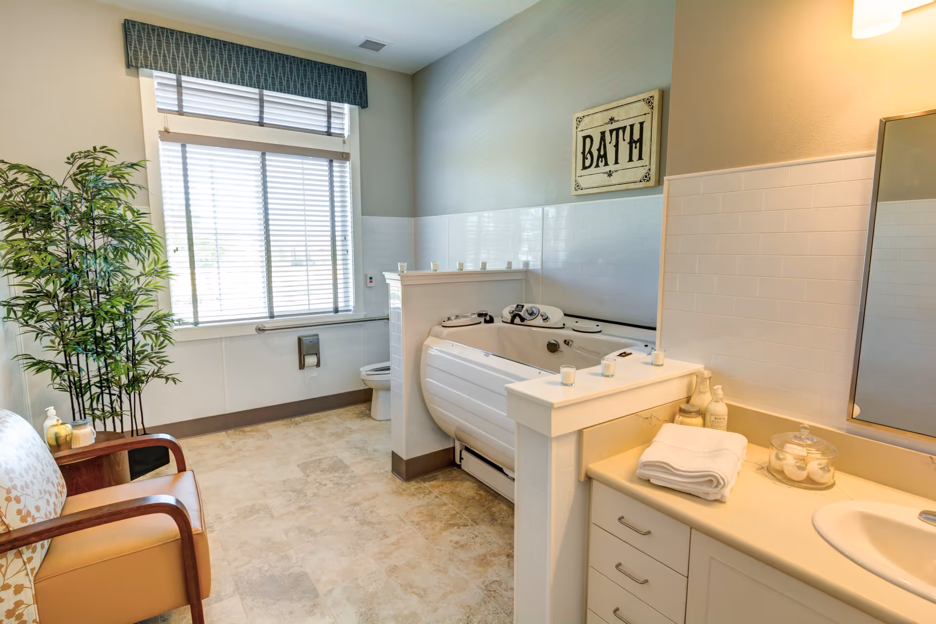 A bright and spacious bathroom with a large window covered by blinds and a valance. The room features a white bathtub with controls, a toilet partially visible behind a half wall, a sink with a countertop holding folded towels and toiletries, a cushioned chair with wooden arms, and a tall green plant in the corner. A decorative sign reading 'BATH' hangs on the wall above the bathtub.