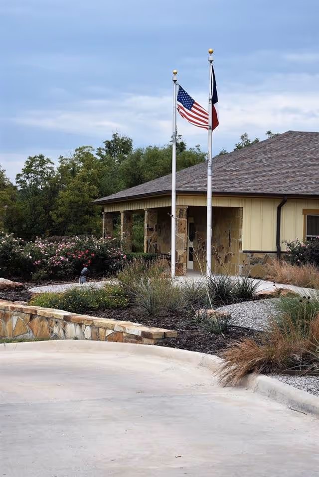 Exterior view of a building with a stone and beige facade, two flagpoles displaying the American flag and another flag, surrounded by landscaped bushes and plants under a cloudy sky.