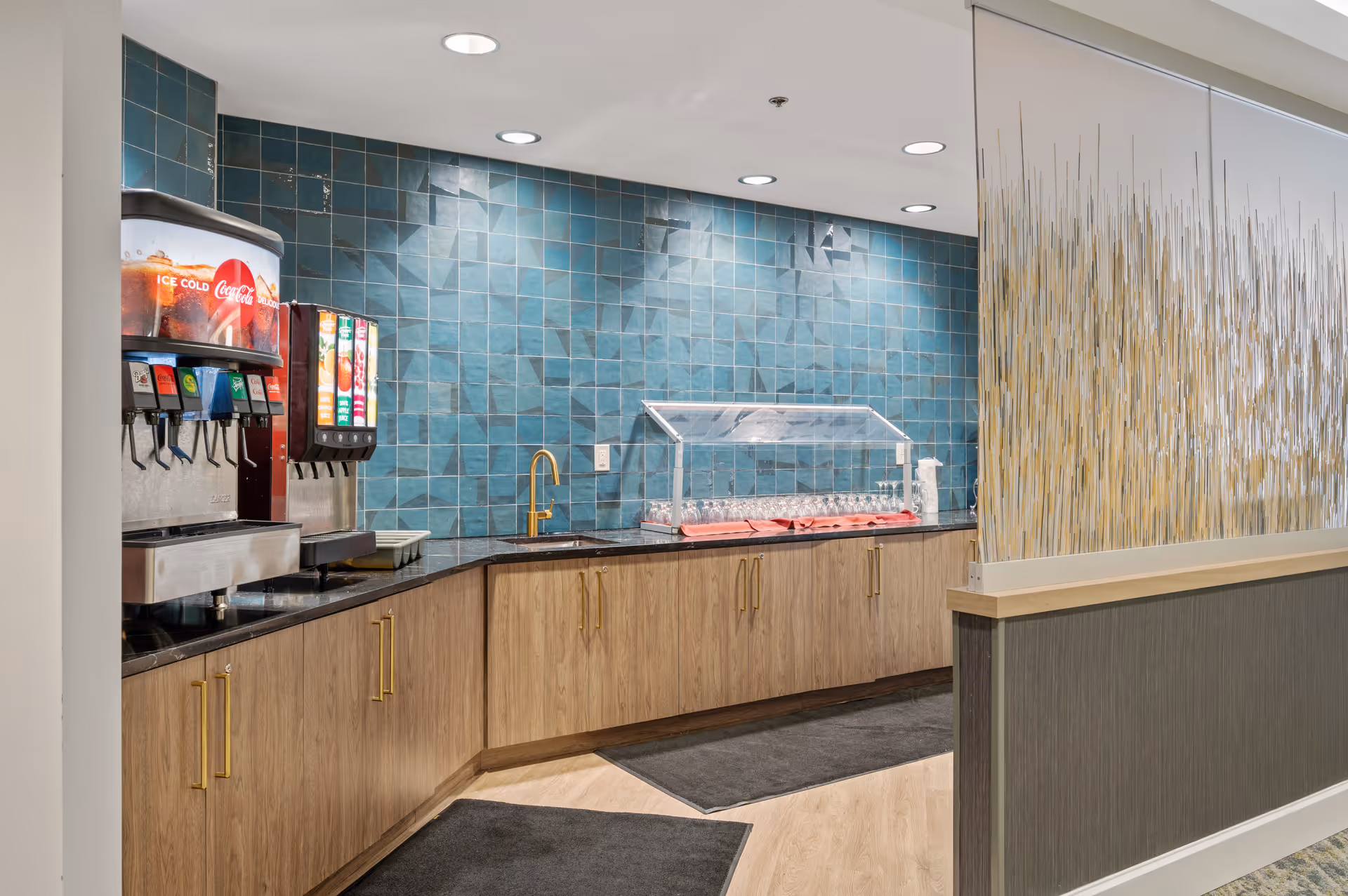 A beverage station with a Coca-Cola soda dispenser, a juice dispenser, and a counter with a glass cover containing glasses. The wall behind the counter is covered with blue-green tiles, and there are wooden cabinets with gold handles below the counter. The floor has light wood and dark mats, and a decorative partition with vertical elements is visible on the right side.