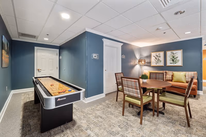 A cozy interior room with a shuffleboard table on the left and a round wooden table with four chairs on the right. Behind the table is a cushioned bench with plaid upholstery and a green pillow. The walls are painted blue with white trim, and two framed botanical prints hang above the bench. A floor lamp with a woven shade provides warm lighting in the corner.