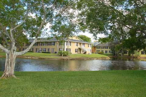 Two-story beige senior living building across a pond with green lawn and trees