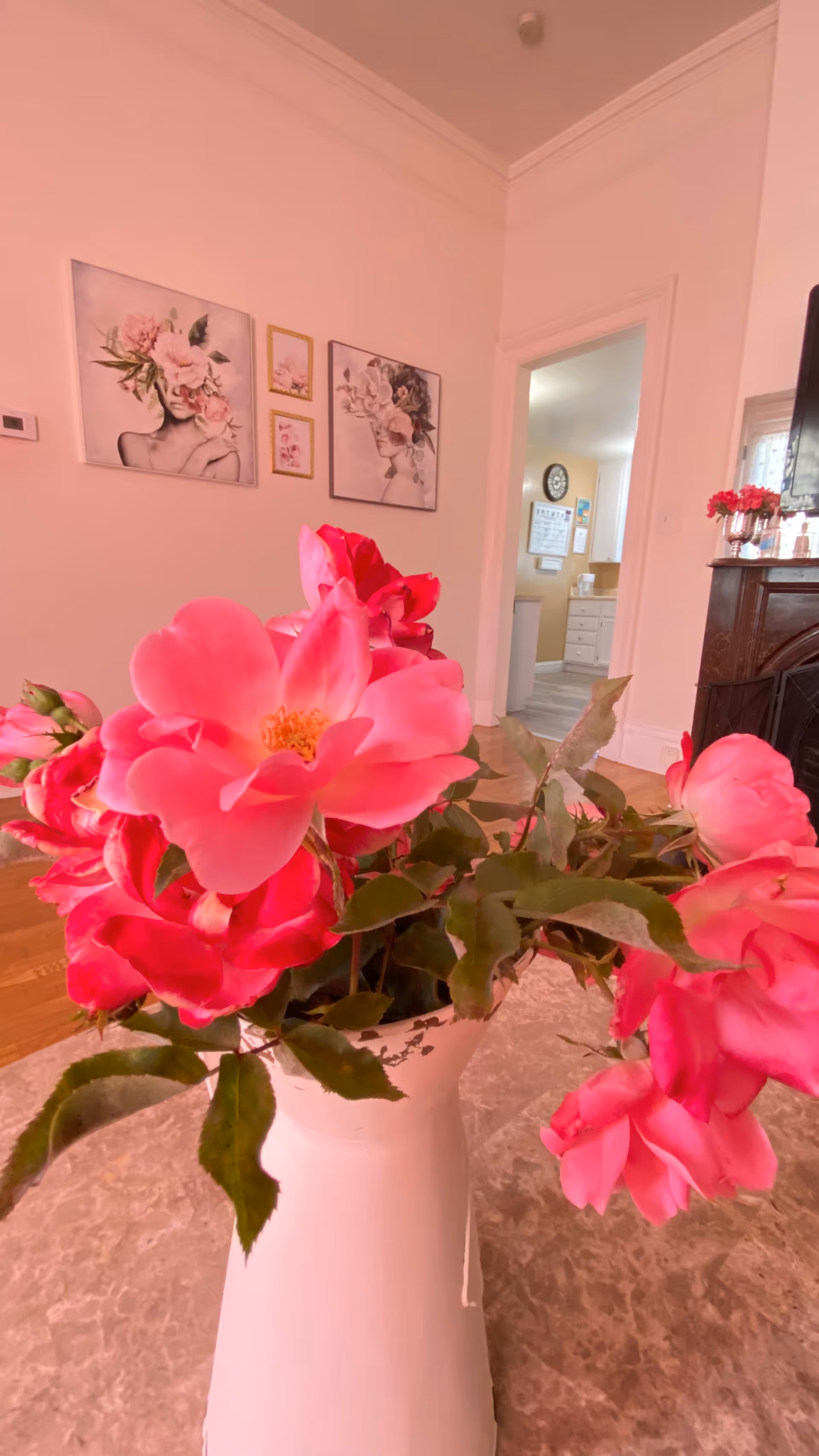 A close-up view of a white vase filled with vibrant pink flowers placed on a marble table in a softly lit room. In the background, there are three framed floral-themed artworks on a light pink wall, an open doorway leading to a kitchen area with white cabinets, and part of a dark wooden fireplace mantel with decorative items on top.